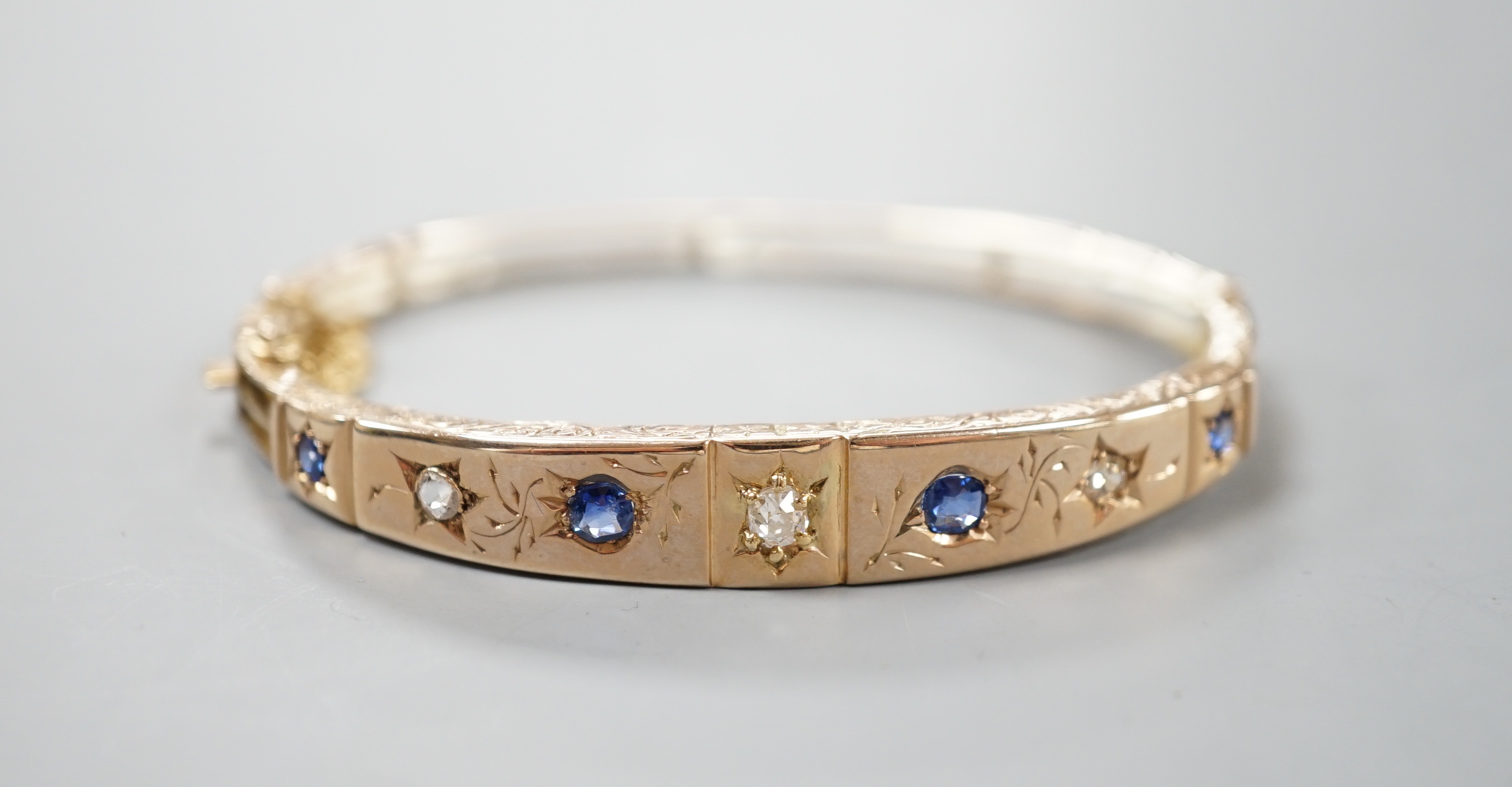 A late Victorian engraved yellow metal, four stone sapphire and three stone diamond set hinged bangle, gross 10.5 grams.